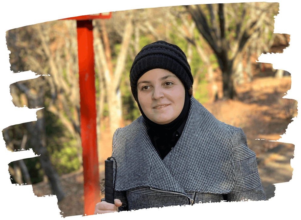 A picture of Mona Minkara smiling for the camera during a sunny day in Japan. She is wearing a grey coat and a black wool hat. Behind her is a path and trees.